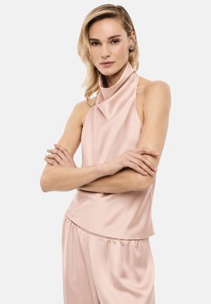 Donna con capelli biondi indossa un top smanicato in raso rosa pallido e pantaloni abbinati, con le braccia incrociate e un’espressione neutra su sfondo bianco.