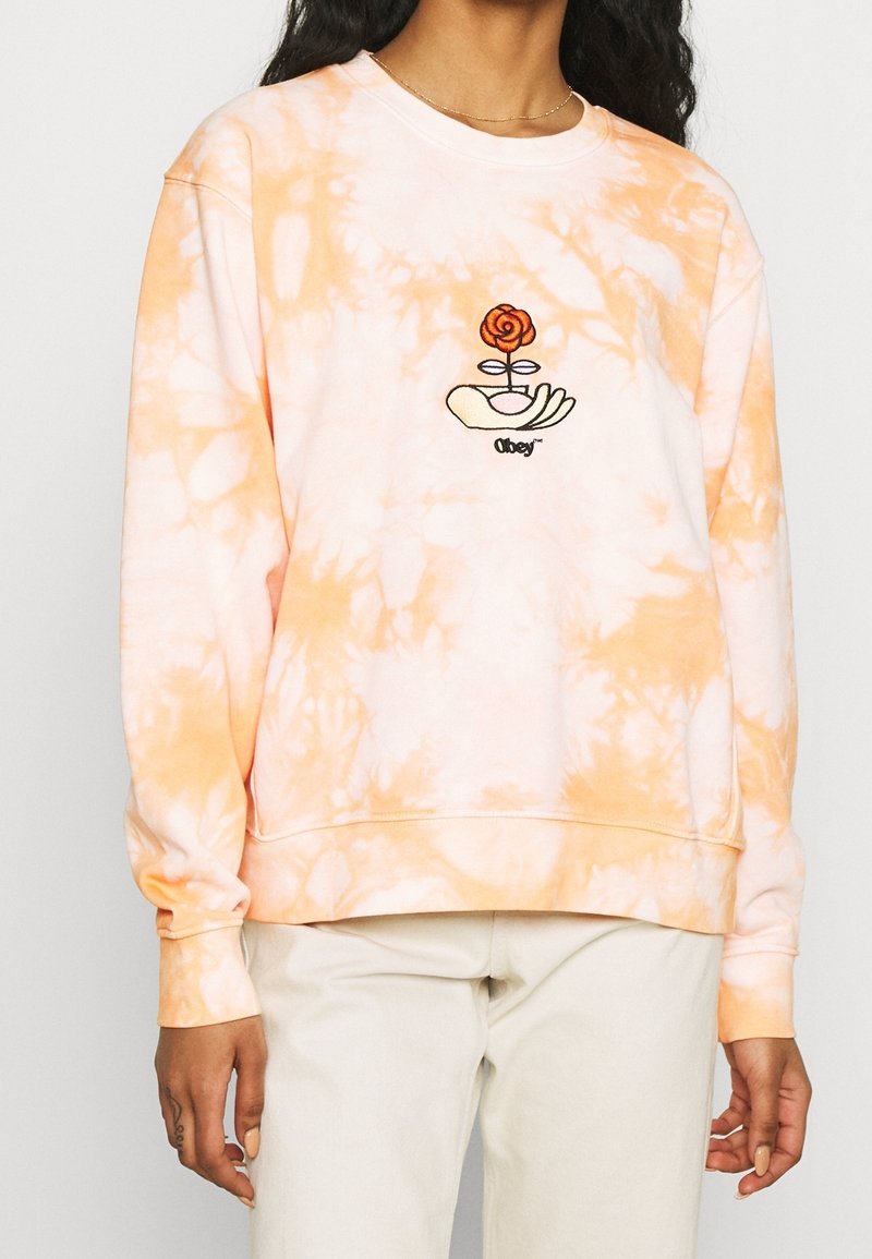 Personne portant un sweat-shirt tie-dye pêche et blanc avec une main brodée tenant une rose et le mot "obey" sur la poitrine.
