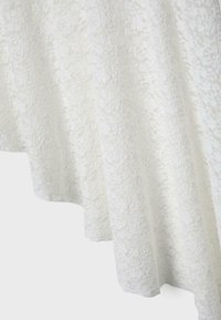 Biały materiał z misterną fakturą kwiatowej koronki i wykończeniem z falistym brzegiem, pokazany z bliska.