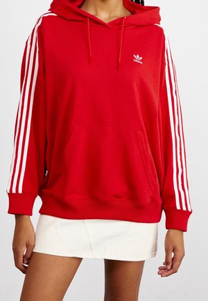 Rød Adidas hættetrøje med hvide tre striber på ærmerne og logo på brystet, båret af en person med flettet hår og hvid nederdel.