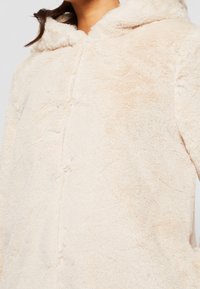 Mujer que lleva una chaqueta de felpa suave color crema con capucha, cuello alto y cierre frontal oculto.