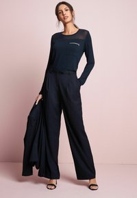 Marineblå langærmet top med gennemsigtige skuldre, brystlomme, parret med højdefaultbukser. Glat tekstur, minimal design. Sorte sandaler og blazer.