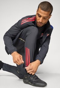 Træningsjakke og bukser i mørkegrå med røde og blå striber; sorte sneakers; model justerer lynlåsen på bukserne ved anklen.
