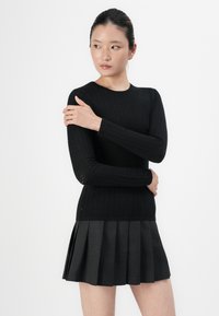 Mlada žena s temnimi lasmi, oblečena v črno teksturirano dolgo-sleeve majico in temno sivo plisirano krilo, stoji z iztegnjenimi rokami.