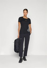 Kobieta w czarnej koszulce z grafiką i czarnych spodniach, trzymająca czarny plecak, stojąca na jednolitym jasnym tle.