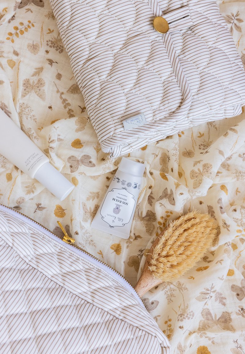 Pochette matelassée avec des rayures crème et beige, accompagnée de produits de soin, d'une brosse en poils naturels et d'un tissu à motif floral.