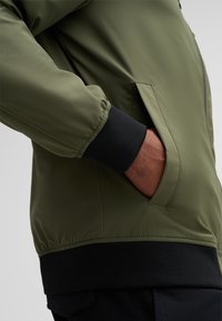 Groene bomberjack met gestructureerde stof, elastische zwarte manchetjes en zijzak. Focus op een gestroomlijnd, modern ontwerp en een getailleerde pasvorm.