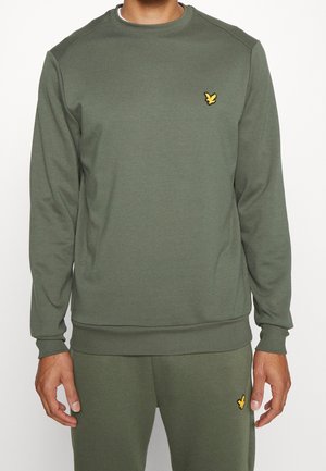 Mand iført olivengrøn sweatshirt og matchende bukser med små gule fuglelogoer på bryst og lår.