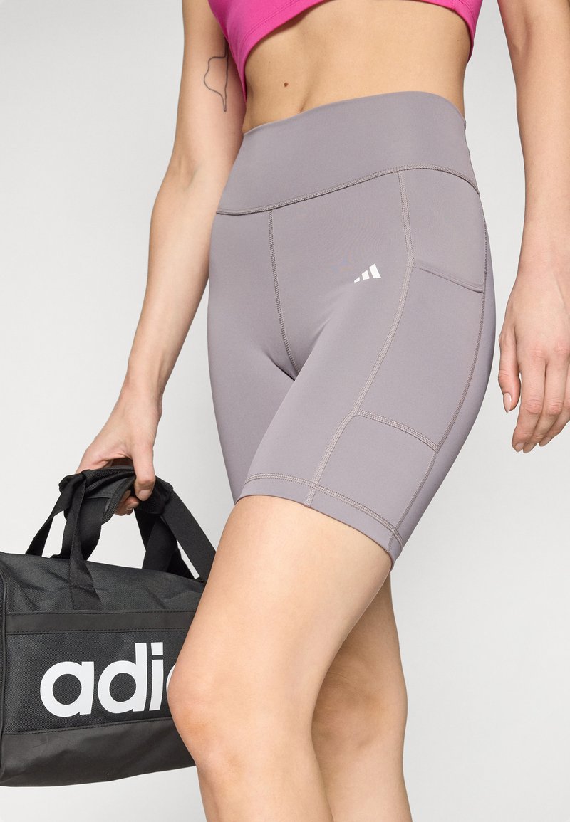 Pantalones cortos deportivos grises con una cinturilla alta, que presentan detalles de costura lateral y un logo de Adidas. El modelo sostiene una bolsa deportiva negra.
