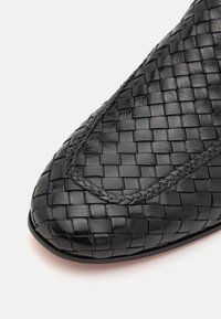 Chaussure en cuir tressé noir avec un bout rond. Présente un tressage intriqué le long de la couture, mettant en valeur une finition texturée. Semelle lisse visible.