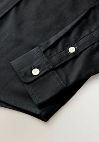 Dettaglio di camicia nera in cotone con polsino abbottonato con bottoni bianchi, cuciture ordinate e una trama liscia.