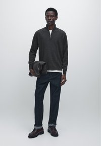 Tmavě šedý svetr na zip s límcem, bílá vnitřní vrstva, tmavě modré džíny, hnědé šněrovací boty, držící šedou bundu. Jednoduchý a minimalistický design.