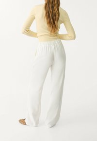 Witte wijdlopende broek van een gladde stof met een elastische tailleband. Gecombineerd met een beige geribbelde top met lange mouwen en bruine slip-on schoenen met studs.