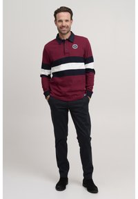 Maroon long-sleeve polo shirt with white and navy horizontal stripes, featuring a collar, button placket, and logo patch. Paired with dark trousers.