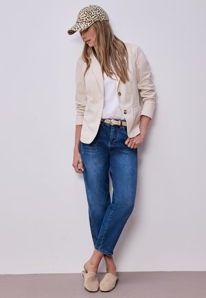 Frau mit Leopardenmuster-Mütze, beigem Blazer, weißem Hemd, blauen Jeans mit Gürtel und beigen Slippers, die vor einer hellgrauen Wand steht.