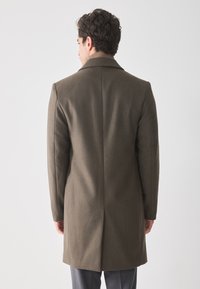 Hombre vestido con un abrigo largo de lana marrón y pantalones grises, de pie de espaldas contra un fondo claro y liso.