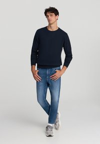 Felpa navy a maniche lunghe, collo rotondo, abbinata a jeans azzurri chiari. Il modello sta in piedi con una mano in tasca, indossando scarpe da ginnastica grigie.