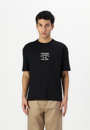 T-shirt en coton noir à manches courtes, avec un texte imprimé blanc comprenant "GUESS", "CALIFORNIA" et "Est. 1981." Design simple.
