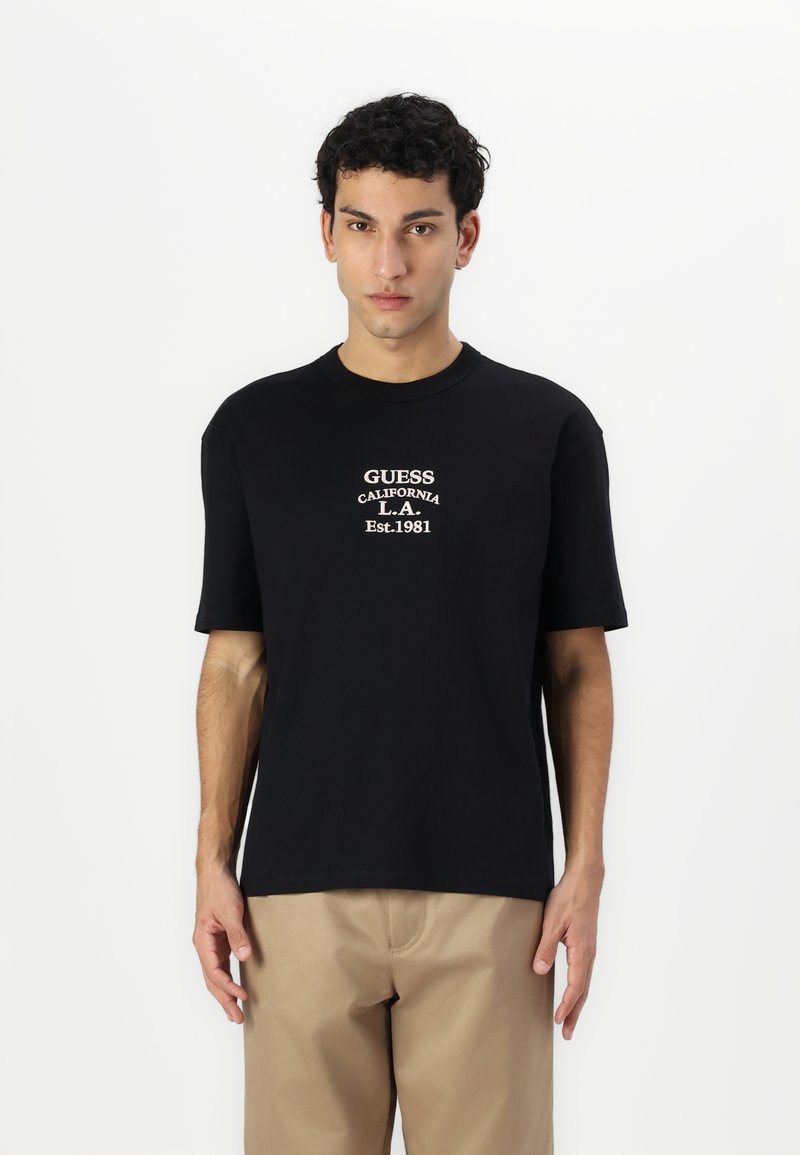 Sort bomulds T-shirt med korte ærmer, der har hvidt trykt tekst, som inkluderer "GUESS," "CALIFORNIA," og "Est. 1981." Simpelt design.