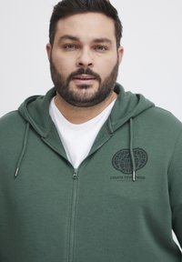 Felpa verde con zip e cappuccio regolabile, caratterizzata da una grafica di un globo nero e testo sul lato sinistro del petto, indossata sopra una maglietta bianca.