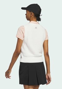 Hvid strikket vest med ribbet kant, båret over en lyserød t-shirt med korte ærmer, parret med en sort plisseret nederdel og en sort kasket.