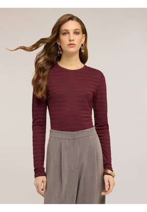 Kvinde med langt, bølget hår iført gyldne øreringe og armbånd, en vinrød stribet langærmet top og højtaljede grå plisserede bukser.