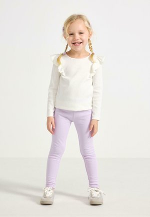 Ung pige med blonde fletninger, smilende, iført en hvid bluse med flæser og lyse lilla leggings, stående mod en hvid baggrund.