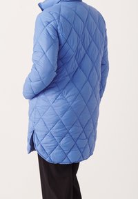 Giacca trapuntata blu con colletto alto e motivo a diamante. Presenta un orlo curvo e spaccature laterali per facilitare il movimento. Sotto ci sono pantaloni neri.