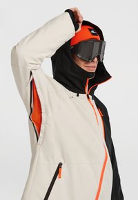 Personne portant une veste d'hiver beige et noire avec une fermeture éclair et des aérations orange, un bonnet orange et des lunettes de ski sombres, soulevant la capuche.