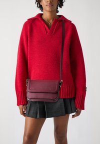 Rød strikket genser med V-hals, kombinert med en vinrød crossover-veske med klaff og smal stropp, båret over et rutete miniskjørt.