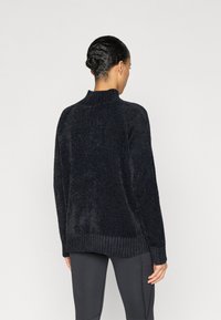 Melns, pūkains adīts džemperis ar augstu apkakli, riboto apakšmali un garām piedurknēm, valkāts virs piegulošām tumšām legingām. Redzama teksturēta virsma.