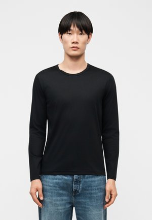 Joven con cabello negro liso que lleva una camisa negra de manga larga lisa y vaqueros azules, posando frente a un fondo gris claro.