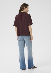 Bordeaux kleurige korte mouwen top met verticale ribbelstructuur en een gerimpelde hals, gecombineerd met lichtblauwe spijkerbroek met flared pijpen. Eenvoudige schoenen zichtbaar.
