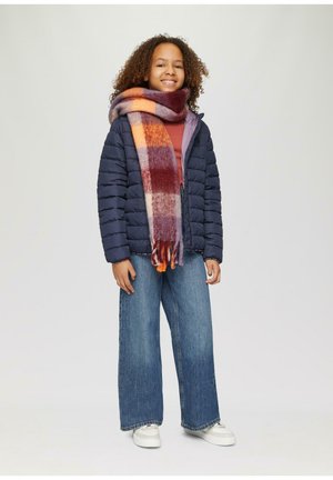 Kind steht, trägt eine marineblaue Steppjacke, weit geschnittene Jeans, weiße Turnschuhe und einen großen karierten Schal in Orange-, Burgunder- und Lilatönen.