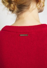 Czerwony sweter z dzianiny z prążkowanym dekoltem. Posiada złotą metkę z logo na plecach. Miękka tekstura, prosty design i żywy kolor.