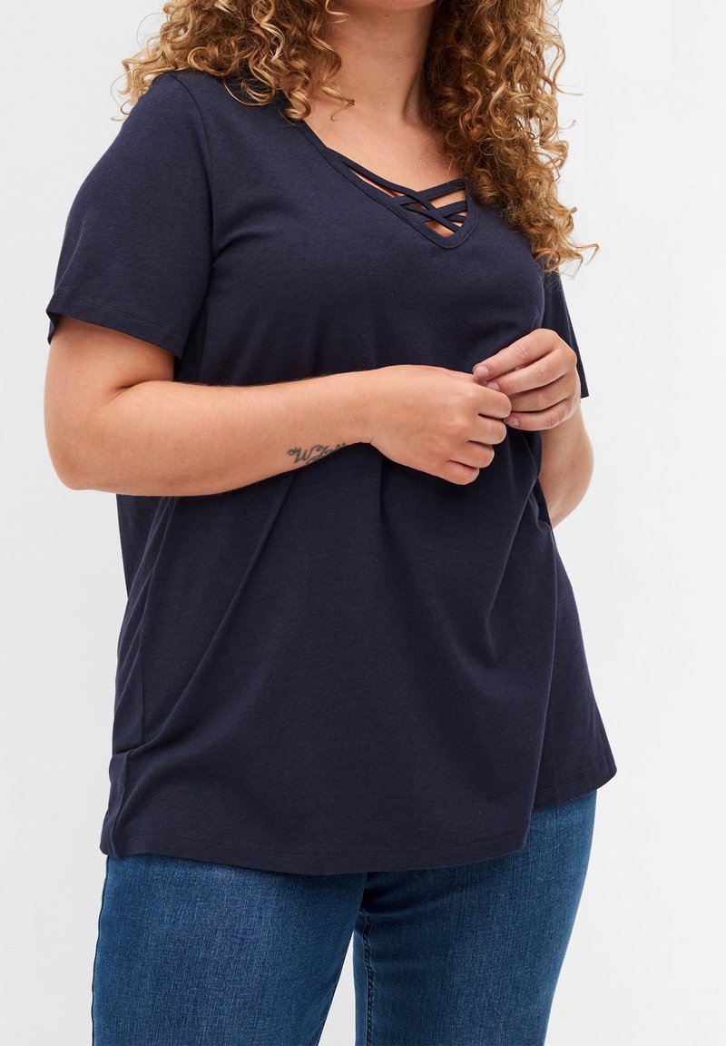 Femme aux cheveux bouclés portant un haut bleu marine à manches courtes avec un décolleté croisé et un jean bleu, mains jointes près de la taille.