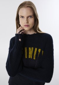 Pull en tricot bleu marine avec un col rond, affichant le texte "INFINI" en jaune. La texture semble douce avec des fils lâches aux poignets.