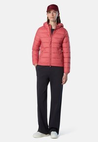 Chaqueta acolchada rosa con capucha, frente con cremallera y bolsillos laterales, combinada con pantalones oscuros y zapatillas blancas. La gorra coincide con el color de la chaqueta.