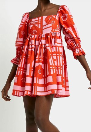 Kobieta ubrana w sukienkę w różowo-czerwony kwiatowy wzór z bufkami o długości 3/4 i kwadratowym dekoltem, stojąca z lekko uniesionymi ramionami.