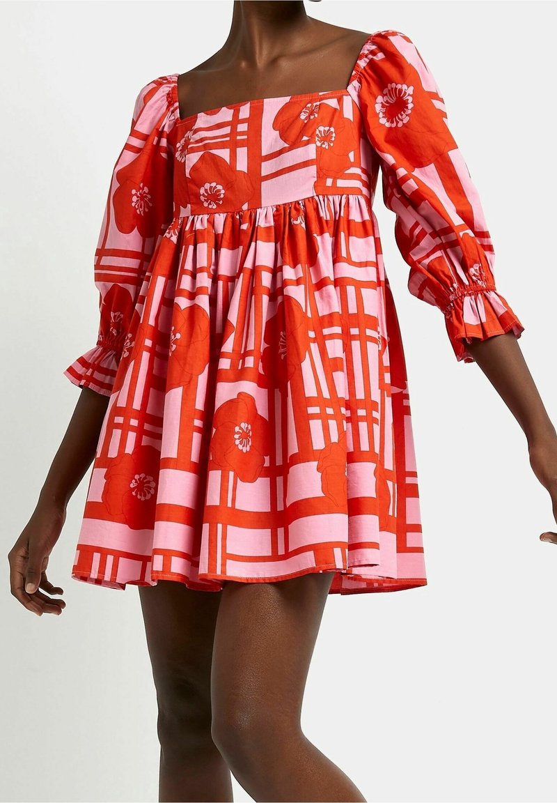 Femme portant une robe à motifs floraux roses et rouges, manches ballon aux trois quarts et encolure carrée, debout avec les bras légèrement levés.