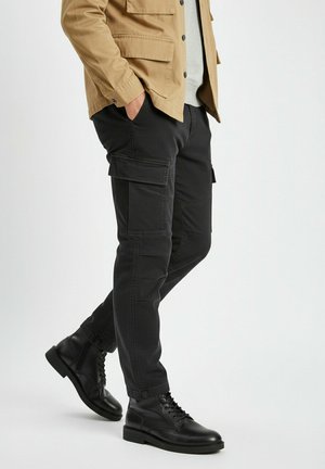 Mann trägt schwarze Cargohosen, schwarze Schnürstiefel, beige Jacke und hellgraues Hemd, steht mit Händen in den Taschen.