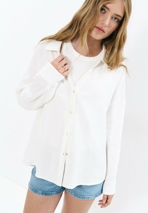 Young woman with long blonde hair wearing a white button-up shirt over a white top and blue denim shorts against a white background.
