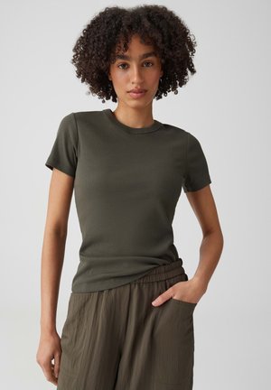 Jeune femme aux cheveux bouclés portant un t-shirt moulant olive à manches courtes et un pantalon ample olive, debout avec une main dans la poche.