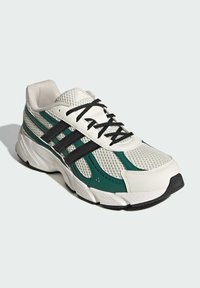 Biela atletická obuv s vrchnou časťou z mastkého materiálu, zelenými a čiernymi akcentmi, čiernymi šnúrkami, textúrovanou podrážkou a tromi čiernymi pruhmi na boku.