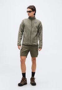 Άνδρας που φοράει πράσινο-ελαιί χνουδωτό μπουφάν, ταιριαστά cargo σορτς, παπούτσια πεζοπορίας, μαύρες κάλτσες και σκούρα γυαλιά ηλίου, στέκεται μπροστά σε απλό φόντο.