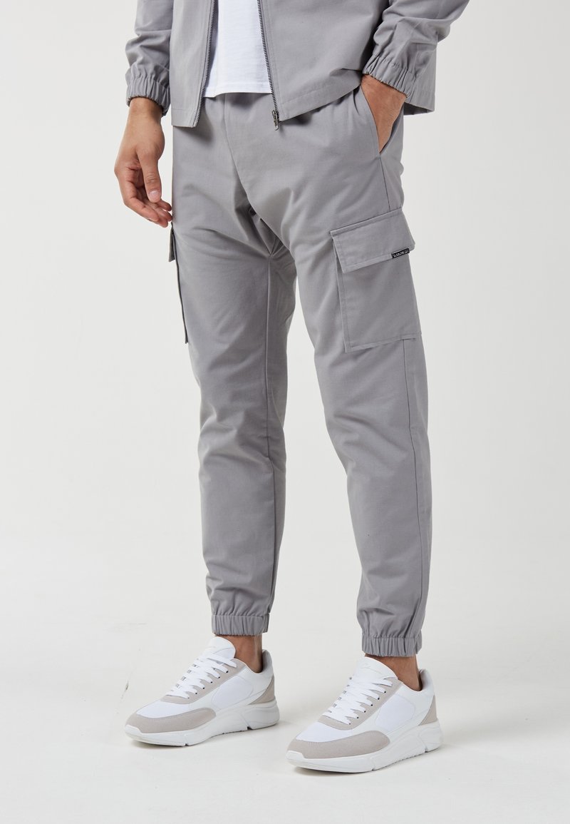 Homme portant un pantalon jogger cargo gris clair et une veste zippée assortie avec des baskets blanches, debout avec une main dans la poche.