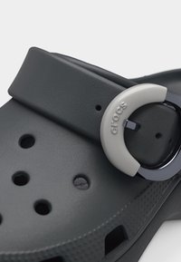 Σκούρο γκρι clog Crocs με κυκλικά οπές αερισμού και ρυθμιζόμενο λουράκι με στρογγυλό ασημί πόμολο χαραγμένο με τη λέξη "crocs".