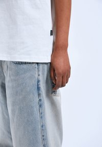 Bílý bavlněný tričko v kombinaci se světle modrými džínovými kalhotami. Mezi jeho vlastnosti patří kapsa na pravé straně a viditelné švy.