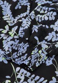 Schwarzer Stoff mit blauen und grünen Blattmustern, ausgestattet mit einem gerafften Bündchen. Das Material wirkt leicht und strukturiert.