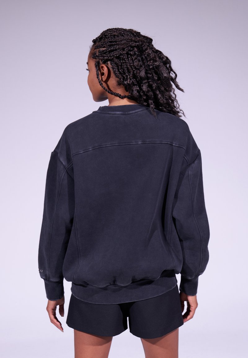 Sudadera negra de gran tamaño con escote redondo, hombros caídos y puños de canalé. Textura suave con detalle de costura visible en la parte posterior.